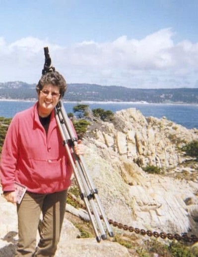 Gael Gallagher stands on the rocky coastline of the Monterey Peninsula, carrying a tripod over her shoulder and a notebook in hand. Dressed in a bright pink jacket, she smiles against the backdrop of rugged cliffs, blue ocean waves, and scenic California beauty.