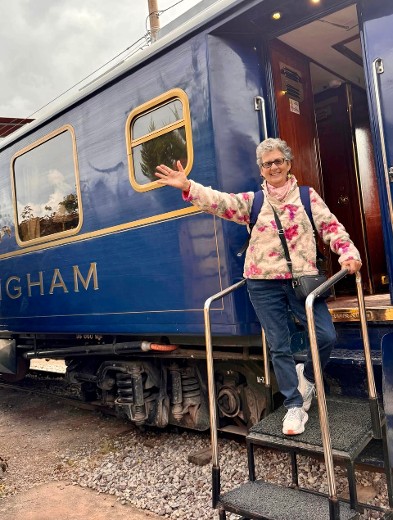  A smiling Gael Gallagher stands on the steps of the luxurious Belmond Hiram Bingham train, dressed casually in jeans and a floral jacket. She waves warmly, ready for an unforgettable journey through Peru’s scenic landscapes.