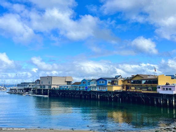  A picturesque view of Fisherman?????s Wharf in Monterey, featuring colorful buildings on stilts over the water, under a bright blue sky.