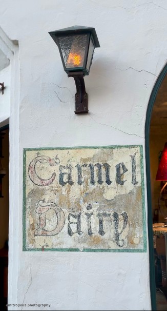 A weathered ‘Carmel Dairy’ sign on a white stucco building, accented by a vintage lantern in Carmel-by-the-Sea.