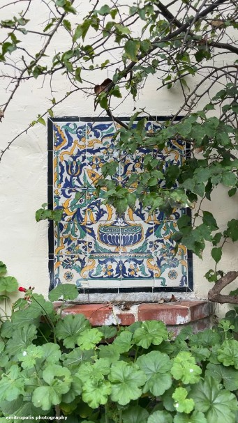 An ornate ceramic tile panel with floral motifs, partially hidden by lush greenery in Carmel-by-the-Sea.