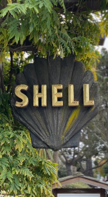 A rustic ‘Shell’ sign shaped like a seashell, nestled among leafy branches in Carmel-by-the-Sea.